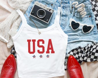 May include: A white tank top featuring the red text USA above three small stars with a red, white, and blue striped pattern. The top is displayed alongside light blue denim shorts, red cowboy boots, heart-shaped sunglasses, and a camera.