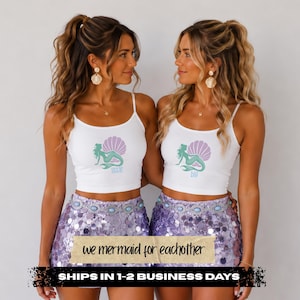 May include: A white tank top featuring a graphic of a green mermaid sitting in a light purple seashell. One top has the word little printed in blue, and the other has the word big printed in blue. Both are paired with a lavender sequin skirt.