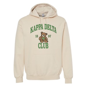 May include: Cream-colored hooded sweatshirt with the text "KAPPA DELTA CLUB" in green, along with a teddy bear graphic. The sweatshirt has a front pocket and a drawstring hood.