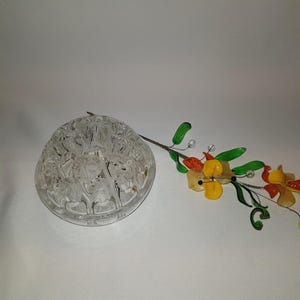 Puede incluir: Un jarrón de flores de vidrio transparente con un diseño decorativo. El jarrón es redondo y tiene una base plana. Un tallo de flor de vidrio con flores de vidrio amarillas, naranjas y verdes está al lado del jarrón.