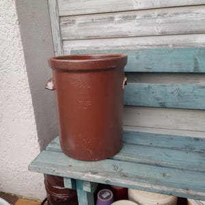 Może przedstawiać: Gliniany garnek w kolorze brązowym o pojemności 2 litrów. Garnek ma dwa małe uchwyty po bokach i stoi na niebieskiej drewnianej ławce.