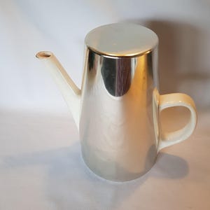 Può includere: Un bricco per la panna in ceramica bianca con corpo argentato e manico. Il bricco ha un beccuccio e una parte superiore arrotondata.