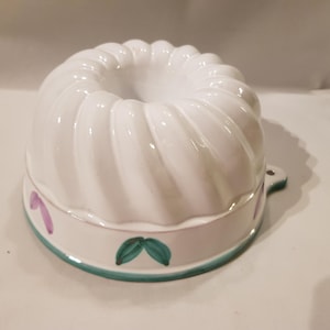 May include: White ceramic bundt cake pan with a fluted design. The pan has a teal rim with painted green leaves and purple accents. A small handle is on the side.