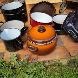 Op de afbeelding: Een verzameling vintage emaille kookgerei in verschillende kleuren, waaronder zwart, rood, oranje en blauw. Het kookgerei omvat potten, pannen en een ovenschaal.