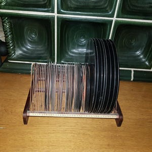 Op de afbeelding: Een bruine houten platenhouder met een verzameling zwarte vinylplaten. De houder heeft een sleufontwerp om de platen te scheiden, met een gedeelte voor het bewaren van een grotere stapel platen.