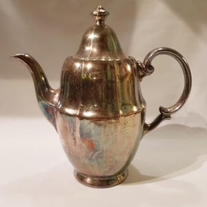 Op de afbeelding: Een zilveren theepot met een rond lichaam en een geribbeld ontwerp. De theepot heeft een deksel met een knop bovenop en een handvat aan de zijkant.