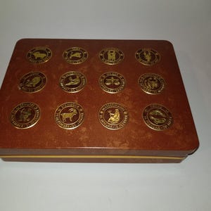 Op de afbeelding: Een bruine houten doos met een gouden rand en een gouden dierenriemsymbool op elk van de 12 vierkanten op de bovenkant van de doos.
