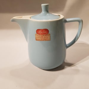 Może przedstawiać: Jasnoniebieski ceramiczny imbryk z pasującą pokrywką i uchwytem. Imbryk ma dzióbek i małą naklejkę z czerwonym i brązowym tekstem. Przedmiot w stylu vintage.
