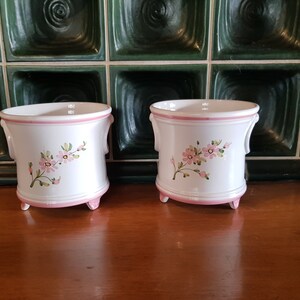 Può includere: Due vasi di fiori in ceramica bianca con bordo rosa e disegni floreali. I vasi hanno tre piccoli piedini e due maniglie sui lati.