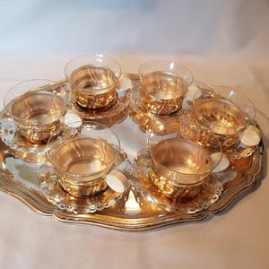 Könnte beinhalten: Ein Vintage-Serviertablett aus versilbertem Metall mit sechs kleinen Gläsern und Untertassen. Die Tassen haben eine goldfarbene Innenseite und stehen auf zarten, spitzenartigen Untertassen. Das Tablett hat einen gewellten Rand.