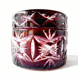 Könnte beinhalten: Eine tiefrote Glasdose mit einem geschliffenen Glasdesign. Die Dose hat ein Sternenmuster und ist aus klarem Glas mit einer roten Tönung gefertigt.
