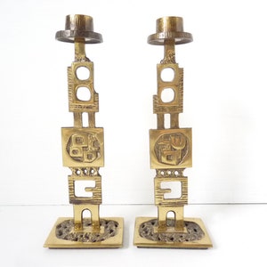 May include: A pair of gold metal candle holders with a geometric design. Each holder has a square base and a tall, slender column with a square top. The columns are decorated with intricate patterns and designs.