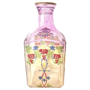 May include: A square glass decanter with a purple hue and a gold top. The decanter is decorated with a floral design and a vine with red grapes.