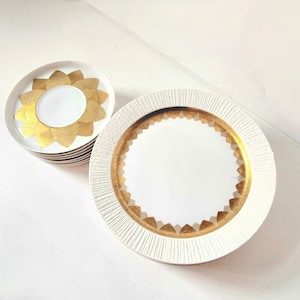 Op de afbeelding: Een set witte keramische borden met gouden accenten. Het grotere bord heeft een getextureerde rand en een gouden band rond de rand. Het kleinere bord heeft een gouden bloemmotief in het midden.