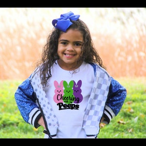 May include: A young girl wearing a white t-shirt with a graphic of three pastel-colored Peeps and the text "Cheering with my Peeps". She is wearing a blue and white checkered jacket and has a blue bow in her hair.