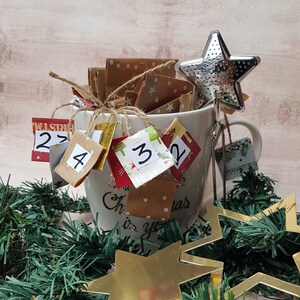 Könnte beinhalten: Eine weiße Keramiktasse mit einem Weihnachts-Adventskalender-Design. Die Tasse hat einen Tee-Infuser am Griff und ist mit kleinen Papierumschlägen gefüllt, auf denen die Zahlen 2, 4, 3 und 2 aufgedruckt sind. Die Tasse ist von grünem Kranz und goldenen Sternen umgeben.