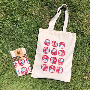 May include: A white tote bag with red and blue daruma dolls printed on it. The bag has two handles and is made of canvas. The bag is sitting on a green grassy surface.