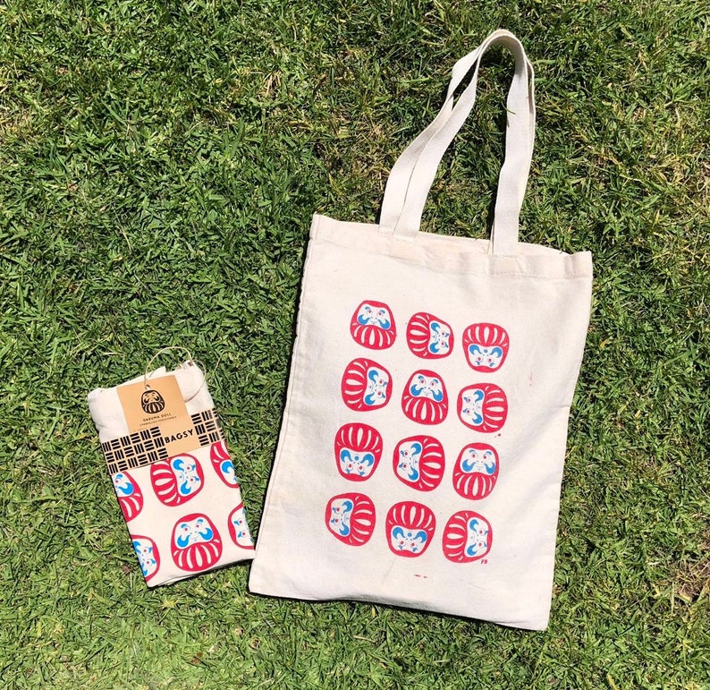 May include: A white tote bag with red and blue daruma dolls printed on it. The bag has two handles and is made of canvas. The bag is sitting on a green grassy surface.
