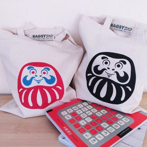 May include: Two white canvas tote bags with red and black daruma doll designs. The bags have the text "BAGSY" printed on them. The bags are sitting on a wooden surface with a red and gray book.