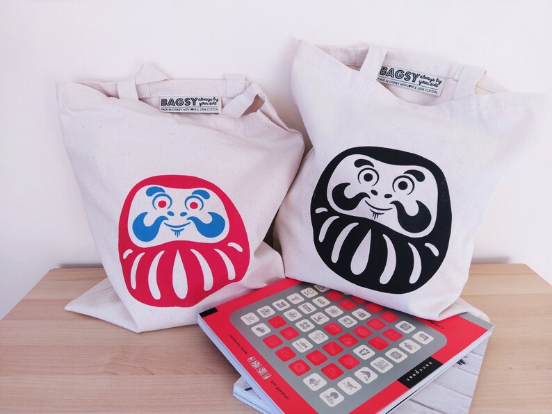 May include: Two white canvas tote bags with red and black daruma doll designs. The bags have the text "BAGSY" printed on them. The bags are sitting on a wooden surface with a red and gray book.