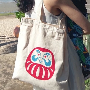May include: A white canvas tote bag with a red and blue daruma doll design. The daruma doll has a smiling face with a mustache and a beard.