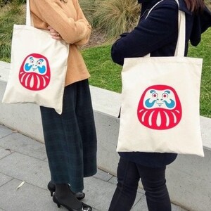 May include: Two white canvas tote bags with red and blue daruma doll designs. The daruma dolls have a smiling face and a red beard. The bags have black handles.