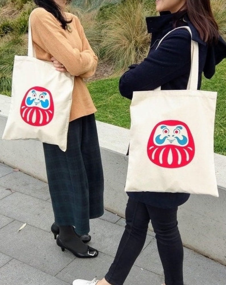 May include: Two white canvas tote bags with red and blue daruma doll designs. The daruma dolls have a smiling face and a red beard. The bags have black handles.