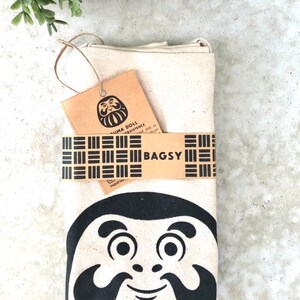 May include: A white canvas tote bag with a black and white illustration of a Daruma doll. The bag has a brown tag with the text "Daruma Doll Persistence" and "Bagsy" printed on it.