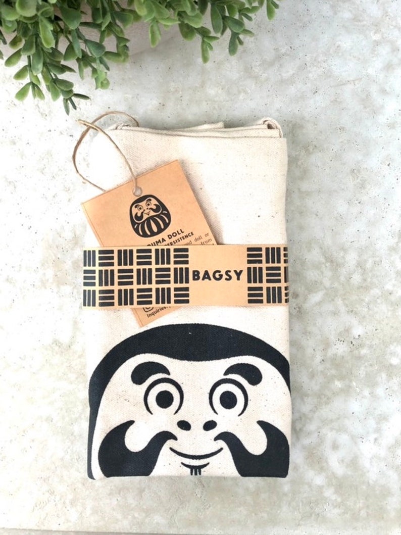 May include: A white canvas tote bag with a black and white illustration of a Daruma doll. The bag has a brown tag with the text "Daruma Doll Persistence" and "Bagsy" printed on it.