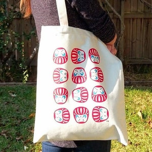 May include: A white tote bag with a red and blue pattern of Japanese Daruma dolls. The bag has a long strap and is being held by a person.