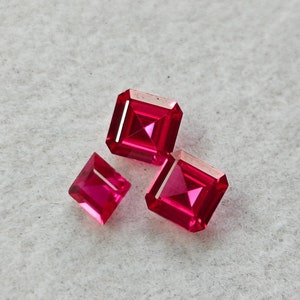 May include: Three red square-shaped gemstones on a white surface.