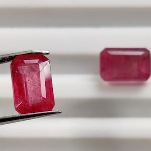 Może przedstawiać: Dwa fasetowane, prostokątne, czerwone kamienie szlachetne. Jeden trzymany jest przez pęsetę, drugi spoczywa na białej powierzchni. Kamienie mają żywy, półprzezroczysty wygląd, ukazujący ich szlif i kolor.