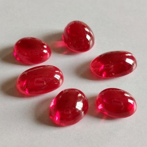 Peut inclure: Six perles de verre rouges translucides de forme ovale. Chaque perle a une surface lisse et polie et un aspect brillant et brillant. Les perles sont dispersées sur une surface blanche, créant un arrangement visuellement attrayant. Idéal pour la fabrication de bijoux ou les projets d'artisanat.