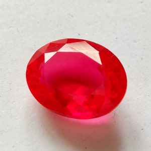 May include: A vibrant, oval-cut gemstone with a deep, saturated red color. The stone features a faceted surface that reflects light, creating a brilliant, translucent appearance with sharp edges and a smooth, polished finish throughout the gem.