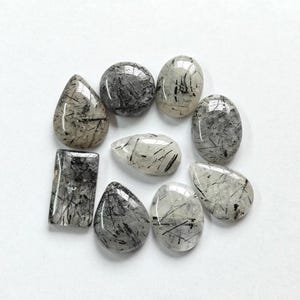 May include: A collection of eight clear quartz cabochons with black inclusions. The cabochons are various shapes and sizes, including oval, teardrop, and rectangular.
