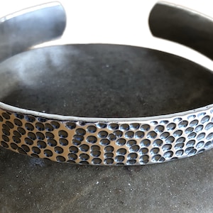 Puede incluir: Un brazalete de plata con textura martillada. El brazalete está hecho de una sola pieza de metal y tiene un borde liso y redondeado.