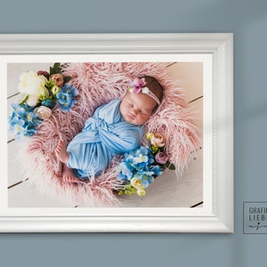 May include: A newborn baby wrapped in a blue blanket is sleeping on a pink fluffy surface surrounded by flowers. The baby is wearing a white headband with a pink flower.