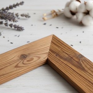 Puede incluir: Primer plano de una esquina de un marco de fotos de madera. El marco es de madera marrón claro con vetas y nudos visibles. El marco está en ángulo, mostrando la unión. Lavanda seca y capullos de algodón en el fondo.