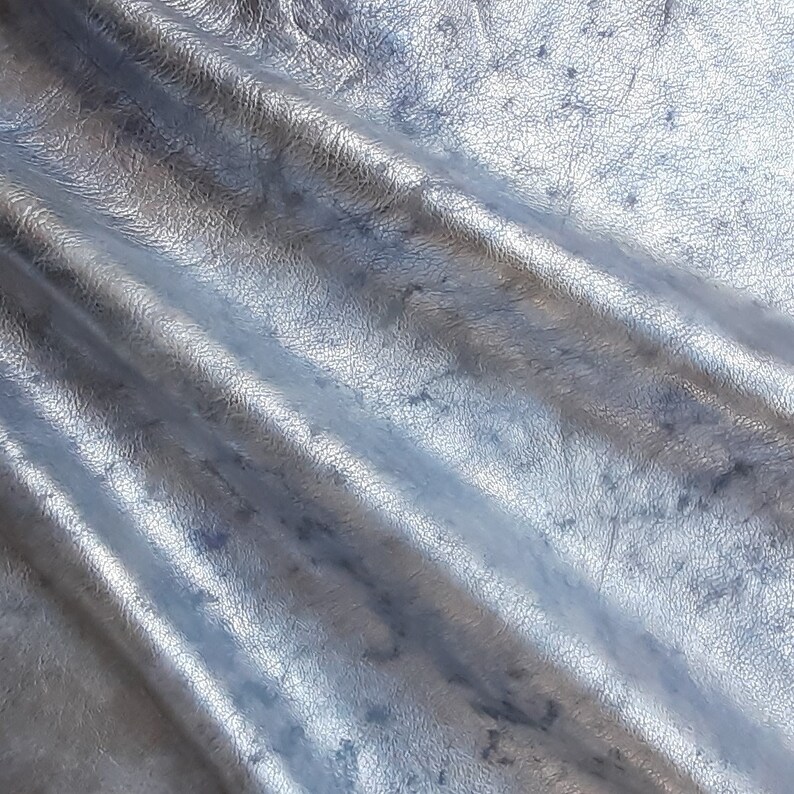 Pu&ograve; includere: Primo piano di un tessuto in pelle argentata con una superficie testurizzata e un aspetto stropicciato.
