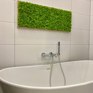 May include: A rectangular piece of wall art with bright green moss in a light brown wooden frame. It's positioned above a white, oval bathtub with chrome fixtures, against a backdrop of white tiled walls. The moss art measures approximately 100cm x 50cm.