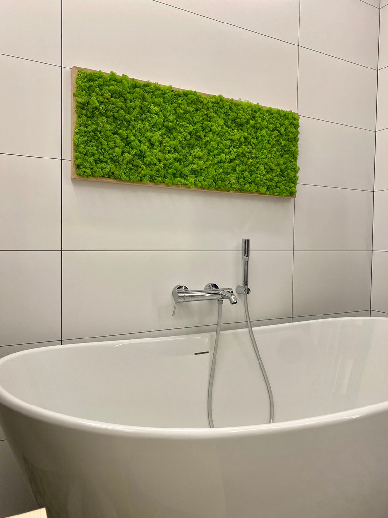 May include: A rectangular piece of wall art with bright green moss in a light brown wooden frame. It's positioned above a white, oval bathtub with chrome fixtures, against a backdrop of white tiled walls. The moss art measures approximately 100cm x 50cm.