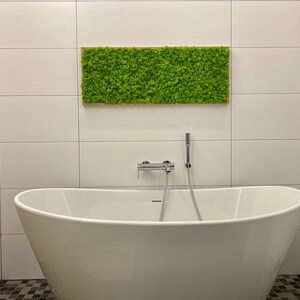 May include: A contemporary bathroom featuring a white, oval freestanding bathtub and a rectangular moss wall art piece. The moss art is framed in wood and hangs above the tub. The walls are white tiled, and the floor has a black and grey mosaic tile.
