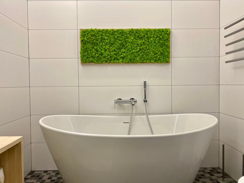 May include: A contemporary bathroom featuring a white, oval freestanding bathtub and a rectangular moss wall art piece. The moss art is framed in wood and hangs above the tub. The walls are white tiled, and the floor has a black and grey mosaic tile.