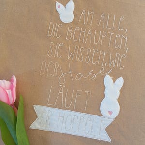 Può includere: Una borsa di tela marrone con testo ricamato bianco che recita "AN ALLC, DIC BCHAUPTCN, SIC WISSEN, WIC DCPflase LÄUFT GR-HOPPCLT". Il testo è circondato da due forme di coniglio in feltro bianco con cuori rosa.