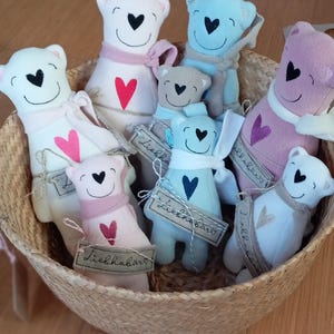 Könnte beinhalten: Eine Sammlung handgefertigter Plüschbären in einem Weidenkorb. Die Bären sind weiß, blau und rosa, jeweils mit einem Herzdetail und einem Schal. Einige Bären haben Etiketten mit der Aufschrift "Liebhabär".