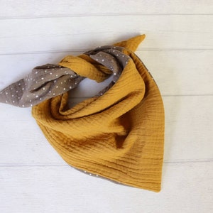 May include: A reversible bandana-style scarf in mustard yellow and brown with white polka dots. The scarf is made of a soft, textured fabric and is knotted at the top. The scarf is displayed on a white wooden surface.