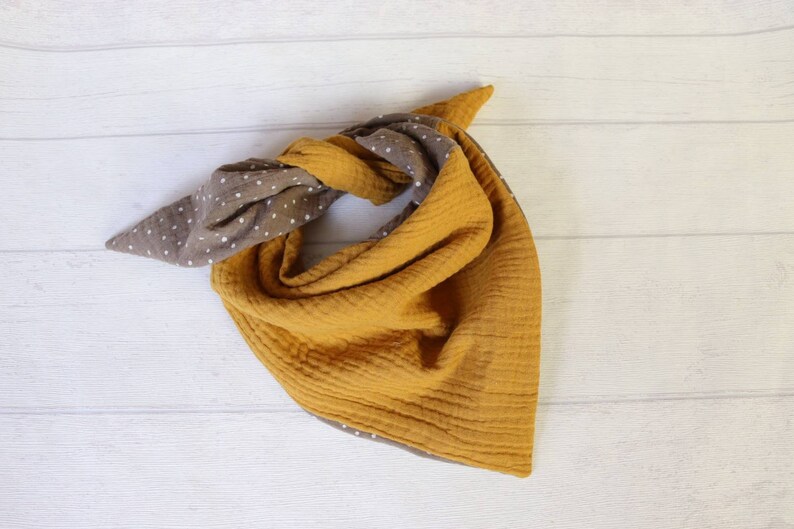 May include: A reversible bandana-style scarf in mustard yellow and brown with white polka dots. The scarf is made of a soft, textured fabric and is knotted at the top. The scarf is displayed on a white wooden surface.