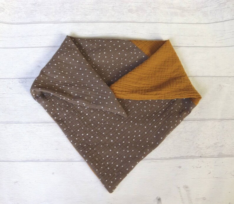 May include: A brown and mustard yellow baby bib. The bib is made of soft, textured fabric. The brown section has a white polka dot pattern. The bib is folded in a triangle shape.
