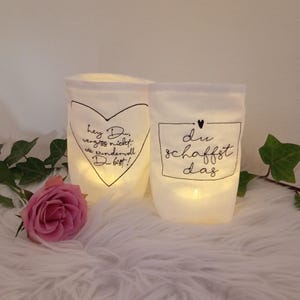 Könnte beinhalten: Zwei weiße Stofflaternen mit beleuchtetem Innenraum. Eine hat einen Herzumriss mit Text, die andere einen quadratischen Umriss mit Text. Eine rosa Rose und Grünzeug befinden sich im Vordergrund. Die Laternen stehen auf einer weißen, flauschigen Oberfläche.