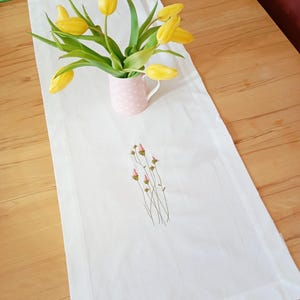 Könnte beinhalten: Weißer Tischläufer mit gestickten rosa Blumen und grünen Stielen. Ein Strauß gelber Tulpen in einem rosa gepunkteten Krug steht auf dem Läufer.